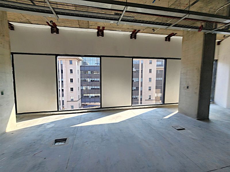 An unfinished open-plan office space featuring exposed concrete ceilings, large windows with urban views, and floor-mounted electrical access points.