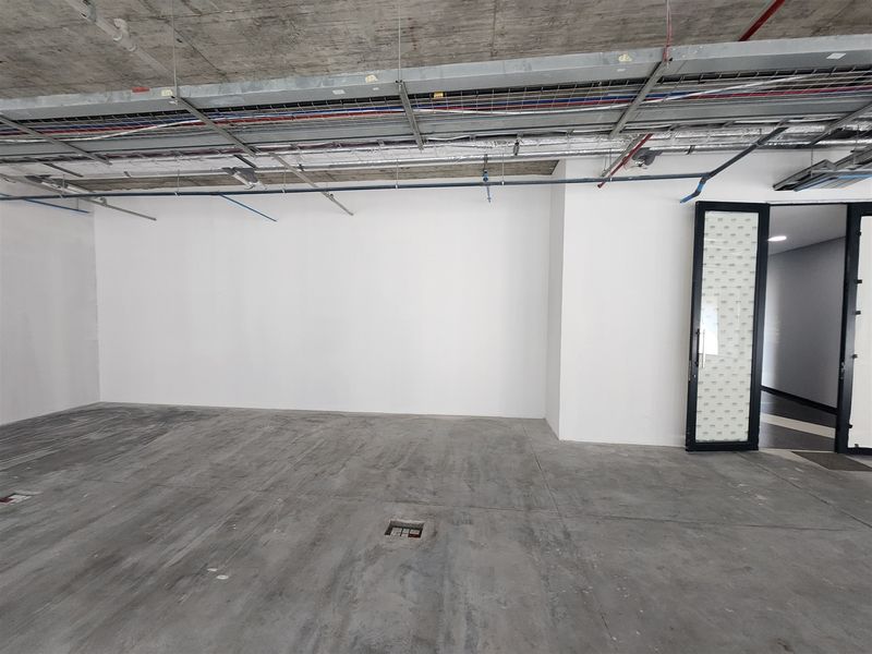 An unfinished open-plan office space featuring exposed concrete ceilings, industrial cable trays, polished concrete flooring, and a modern glass-paneled entrance door.