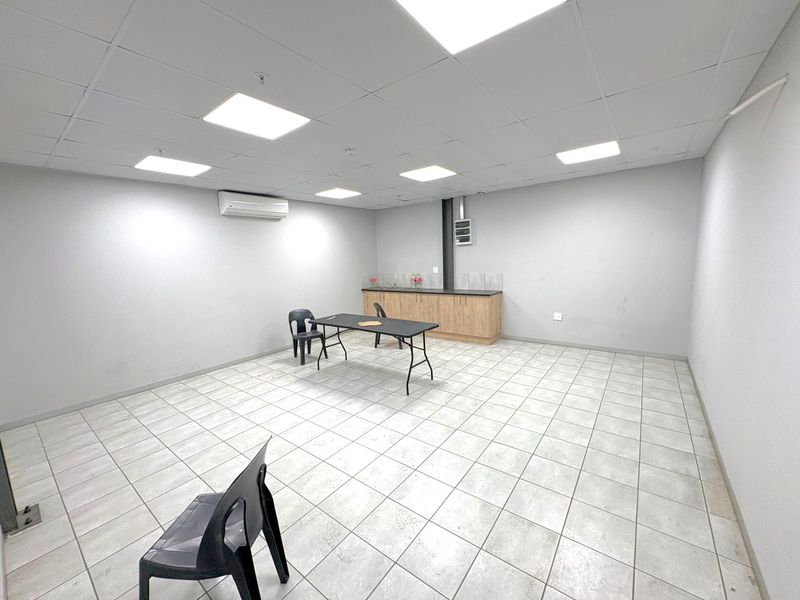 A clean, open-plan office space featuring a suspended ceiling with recessed lighting, tiled floors, a wall-mounted air conditioning unit, and a small kitchenette area.