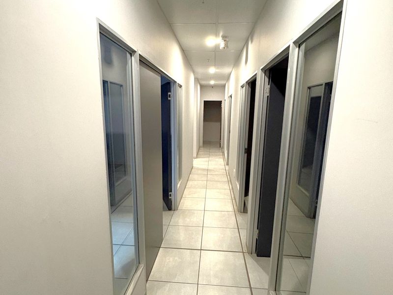 A clean, well-lit industrial hallway featuring tiled flooring, a suspended ceiling with recessed lighting, and multiple glass-panelled office doors.