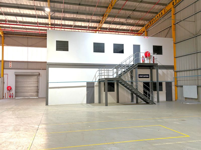Spacious industrial warehouse featuring a mezzanine level with office or canteen space, steel staircase, roller shutter door, and high-clearance exposed roof structure.