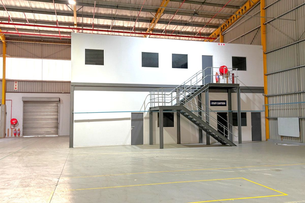 Spacious industrial warehouse featuring a mezzanine level with office or canteen space, steel staircase, roller shutter door, and high-clearance exposed roof structure.