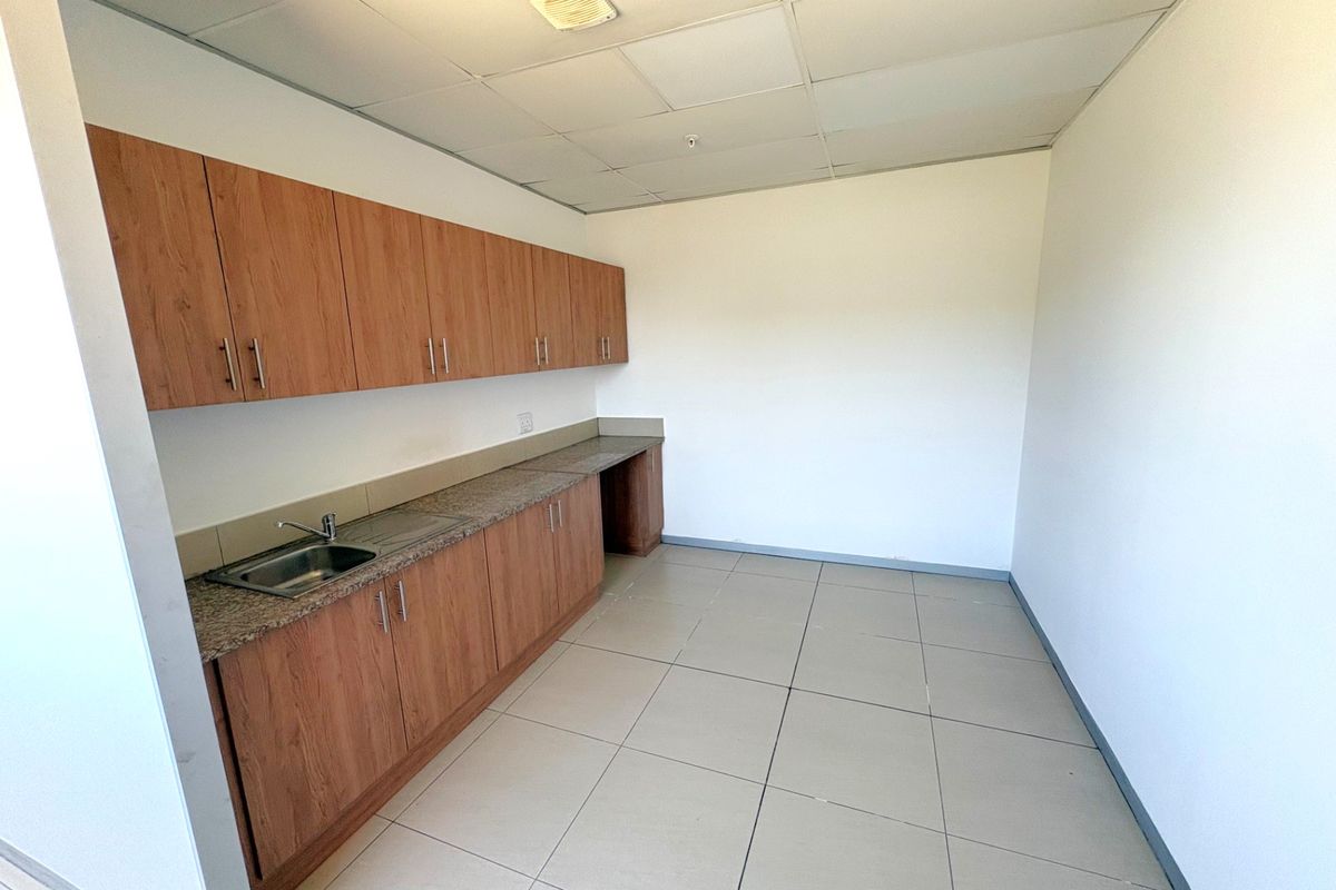 A clean, empty kitchenette area featuring wood-look laminate cabinetry, granite countertops, a stainless steel sink, and neutral ceramic floor tiles.