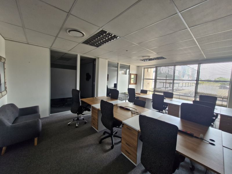 A professional open-plan office space featuring glass partitions, suspended ceilings, carpeted flooring, and large windows providing ample natural light.