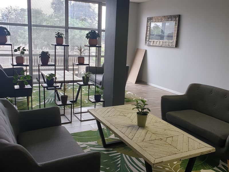 A commercial office lounge area featuring large windows with venetian blinds, a central structural column, and a suspended ceiling with recessed lighting.