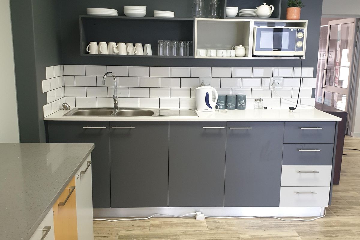 A modern commercial kitchenette featuring dark grey cabinetry, white subway tile backsplash, stone countertops, and a double sink with a mixer tap.
