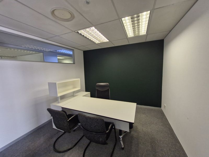 A compact private office space featuring a suspended ceiling with recessed lighting, carpeted flooring, and an internal glass partition wall.