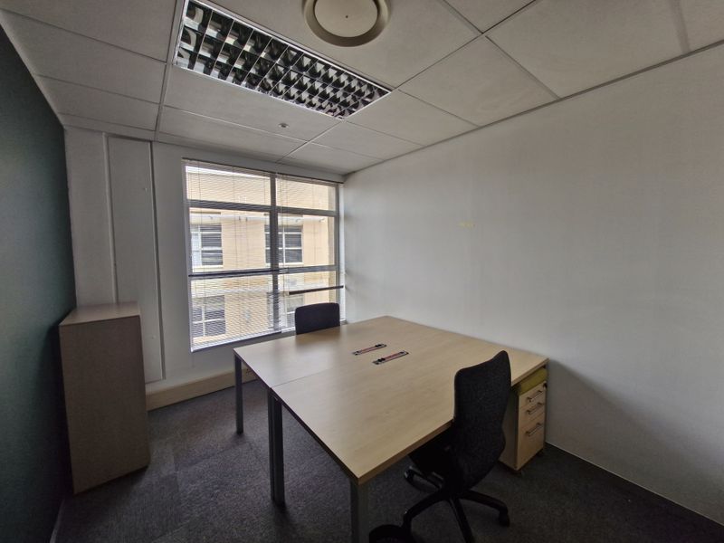 A functional commercial office space featuring carpeted flooring, a suspended ceiling with recessed lighting, and large windows with venetian blinds.