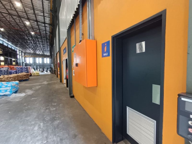 Industrial warehouse interior featuring high ceilings with exposed steel trusses, concrete flooring, and access to designated male restroom facilities.