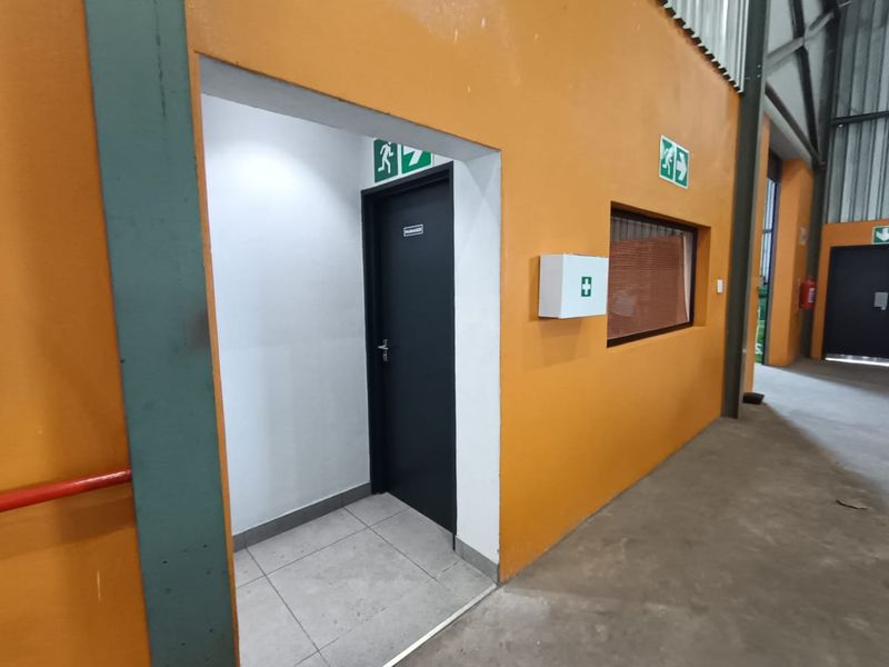 Industrial hallway area featuring tiled flooring, emergency signage, and a wall-mounted first aid kit within a functional warehouse space.