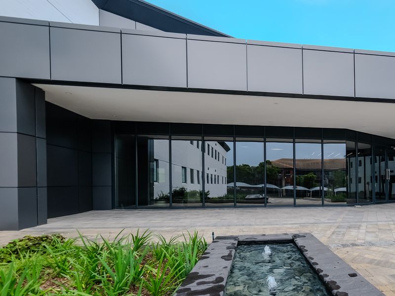 Modern industrial building entrance featuring sleek metal cladding, extensive glass curtain walling, and a decorative stone water feature in the paved forecourt.