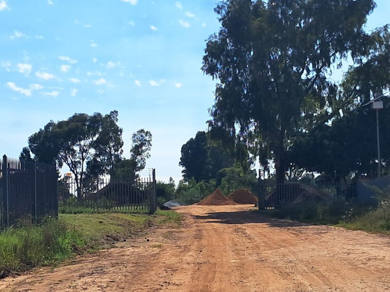View of the commercial vacant land entrance featuring perimeter steel palisade fencing, security gate, and mature trees on a dirt access road.