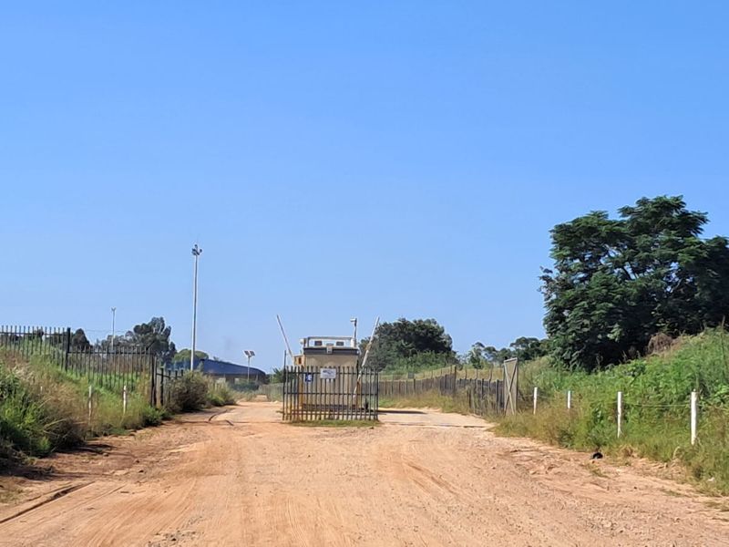 View of a commercial vacant land access point featuring a security gatehouse, perimeter fencing, and an unpaved dirt road entrance.