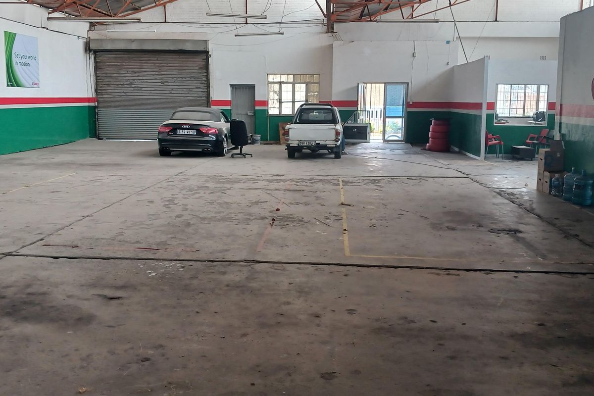 Spacious industrial warehouse featuring high exposed steel truss ceilings, concrete flooring, and a large roller shutter door for vehicle access.