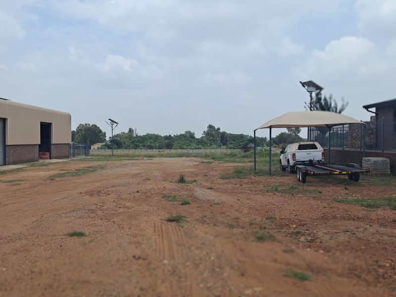 A view of the commercial property grounds featuring a large warehouse structure, a shaded parking area, and open unpaved land.