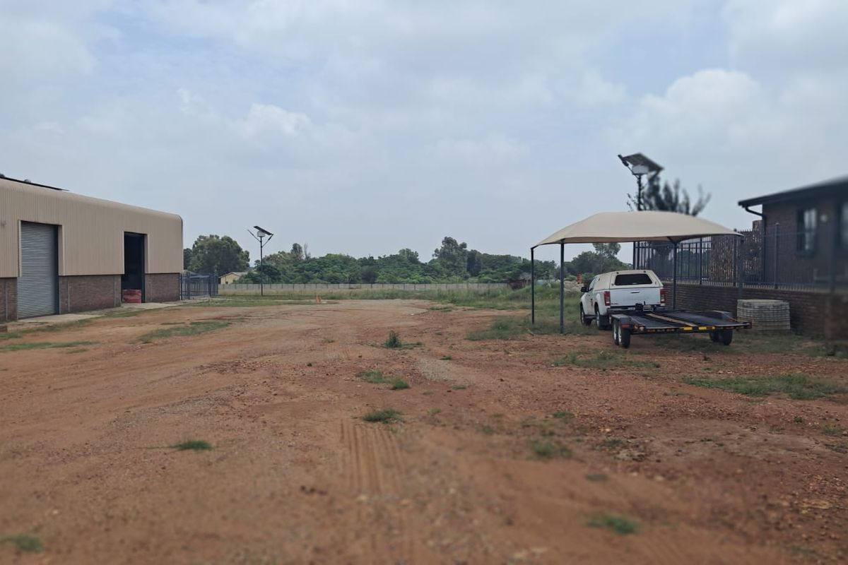 A view of the commercial property grounds featuring a large warehouse structure, a shaded parking area, and open unpaved land.