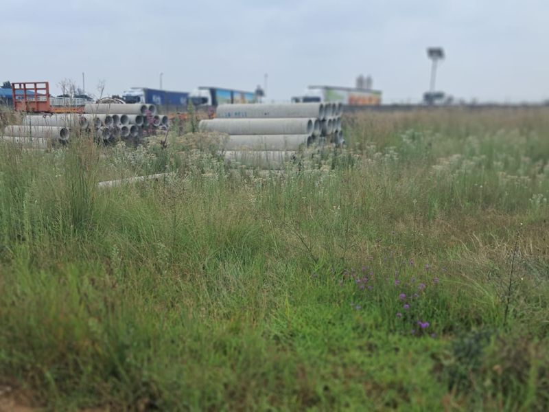 A view of a vacant commercial land plot featuring overgrown grass and stacked concrete drainage pipes on site.