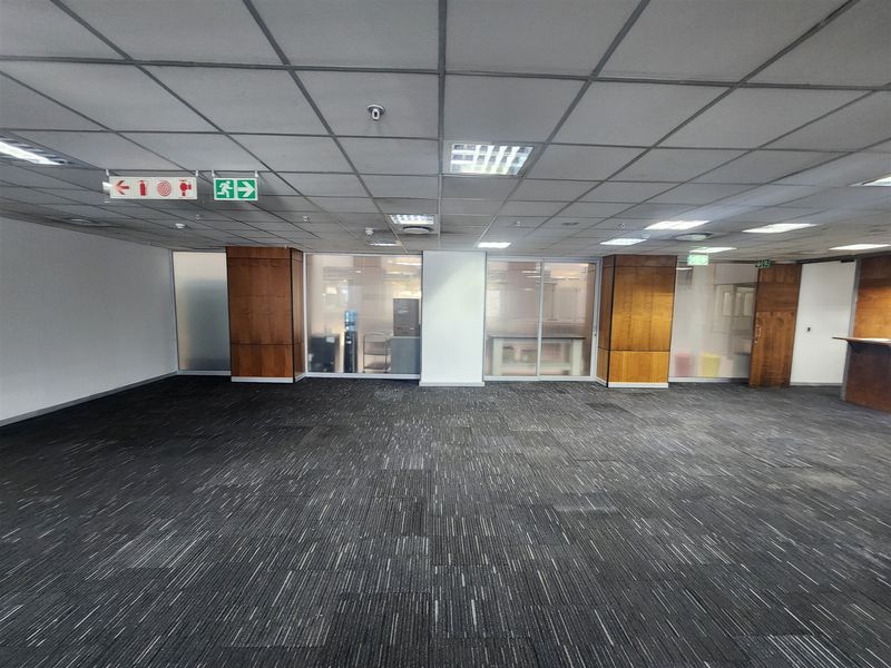 An open-plan office space featuring carpeted flooring, a suspended ceiling with recessed lighting, and glass partitioning with wooden wall accents.
