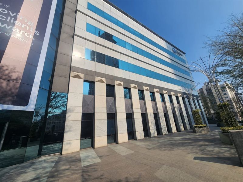 Modern multi-story office building featuring a glass curtain wall facade, stone cladding, and a clean paved pedestrian entrance area.