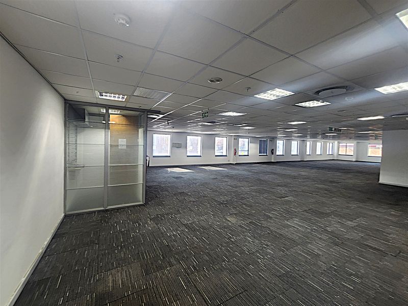 A spacious, open-plan office area featuring a suspended ceiling, recessed lighting, carpet tiles, and a glass-partitioned private office space.