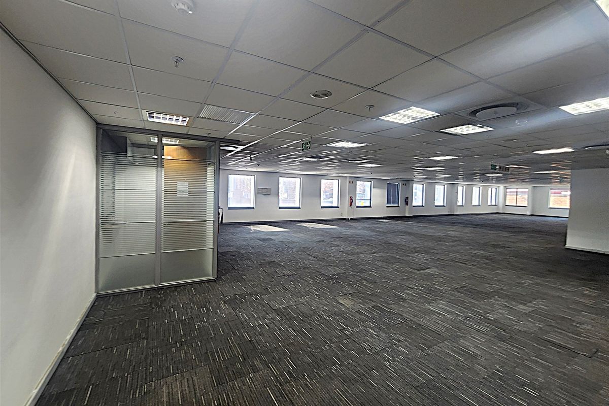 A spacious, open-plan office area featuring a suspended ceiling, recessed lighting, carpet tiles, and a glass-partitioned private office space.