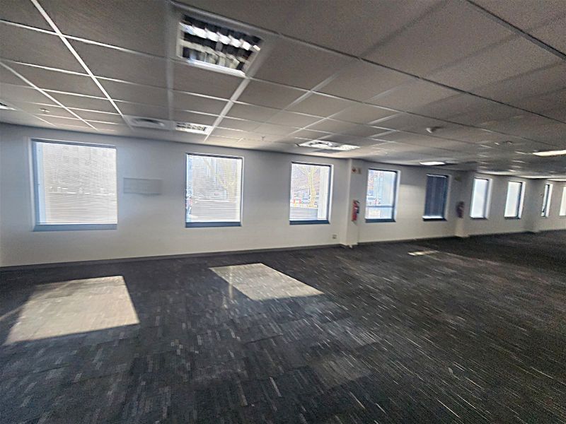 A spacious, open-plan office area featuring suspended ceilings, recessed lighting, and carpet tiles, with large windows providing ample natural light.