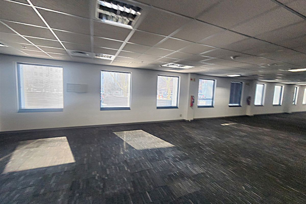 A spacious, open-plan office area featuring suspended ceilings, recessed lighting, and carpet tiles, with large windows providing ample natural light.