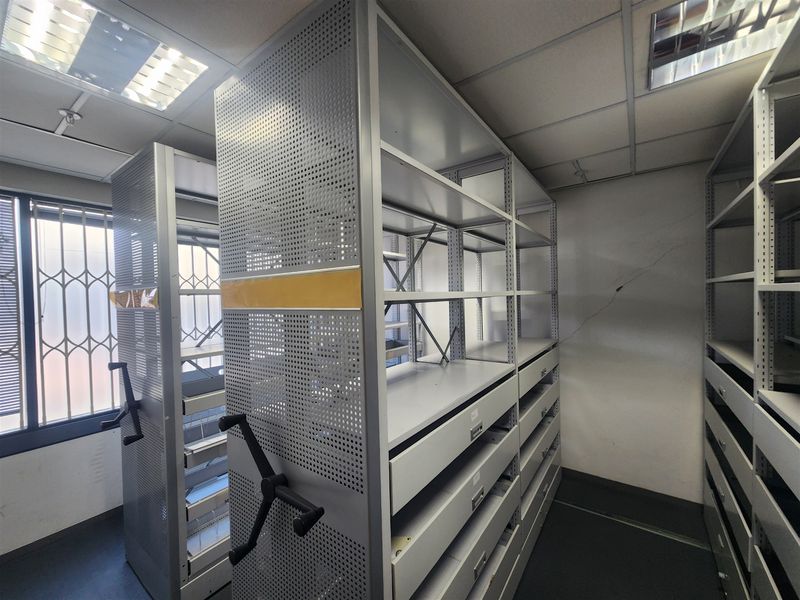 A utility storage room featuring industrial mobile shelving units, suspended ceiling with recessed lighting, and security bars on the windows.