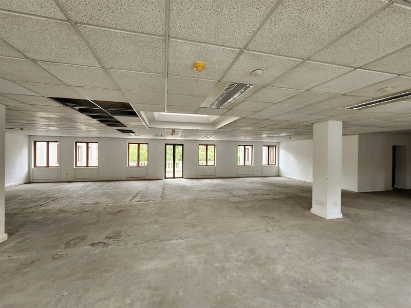 A large, open-plan office space featuring a suspended ceiling, recessed lighting, a central structural column, and expansive windows providing natural light.