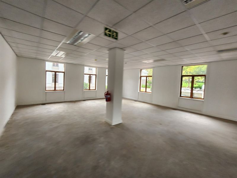 A spacious, vacant open-plan office area featuring a suspended ceiling, recessed lighting, and large windows providing ample natural light.