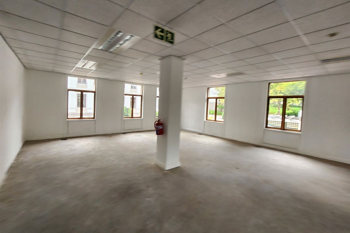 A spacious, vacant open-plan office area featuring a suspended ceiling, recessed lighting, and large windows providing ample natural light.