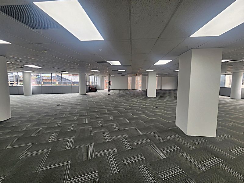 Spacious open-plan office area featuring carpet tiles, suspended ceilings with recessed lighting, and large windows providing ample natural light.