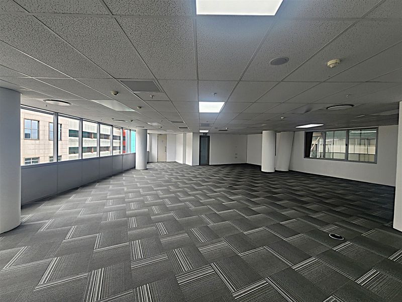 A spacious, open-plan office area featuring modern carpet tiles, a suspended ceiling with recessed lighting, and large windows providing ample natural light.