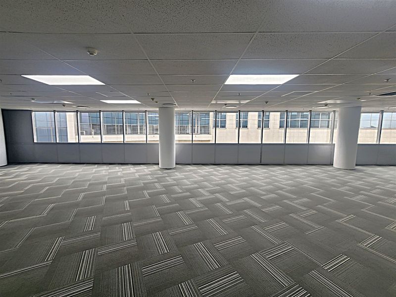 A spacious, open-plan office area featuring carpeted flooring, structural columns, and large windows providing ample natural light.