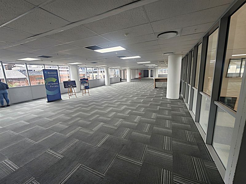 Spacious open-plan office area featuring large windows, carpeted flooring, suspended ceilings with recessed lighting, and glass partitioning for a bright, professional workspace.