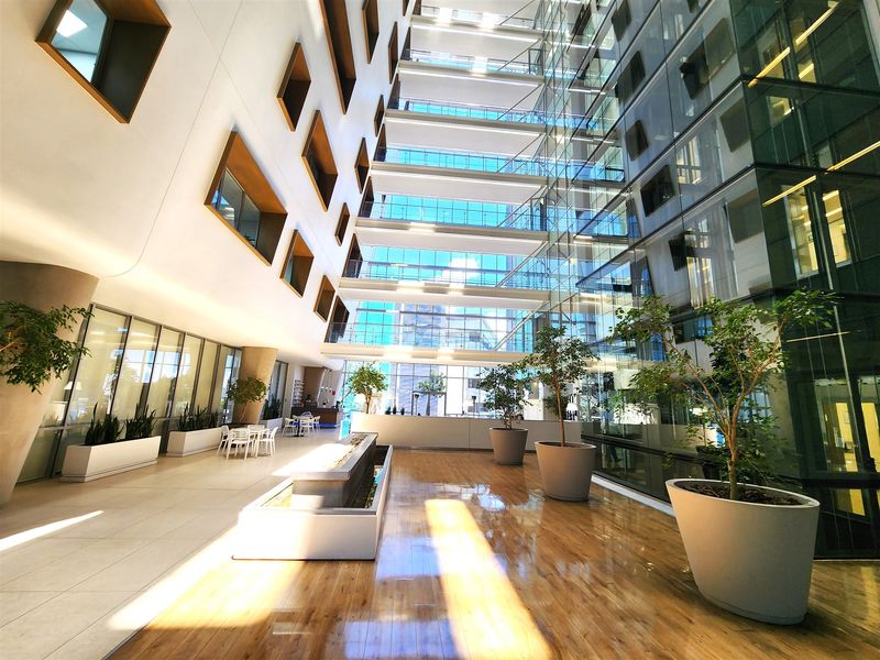A modern, spacious office atrium featuring a multi-story glass facade, polished hardwood and tile flooring, and a central water feature.