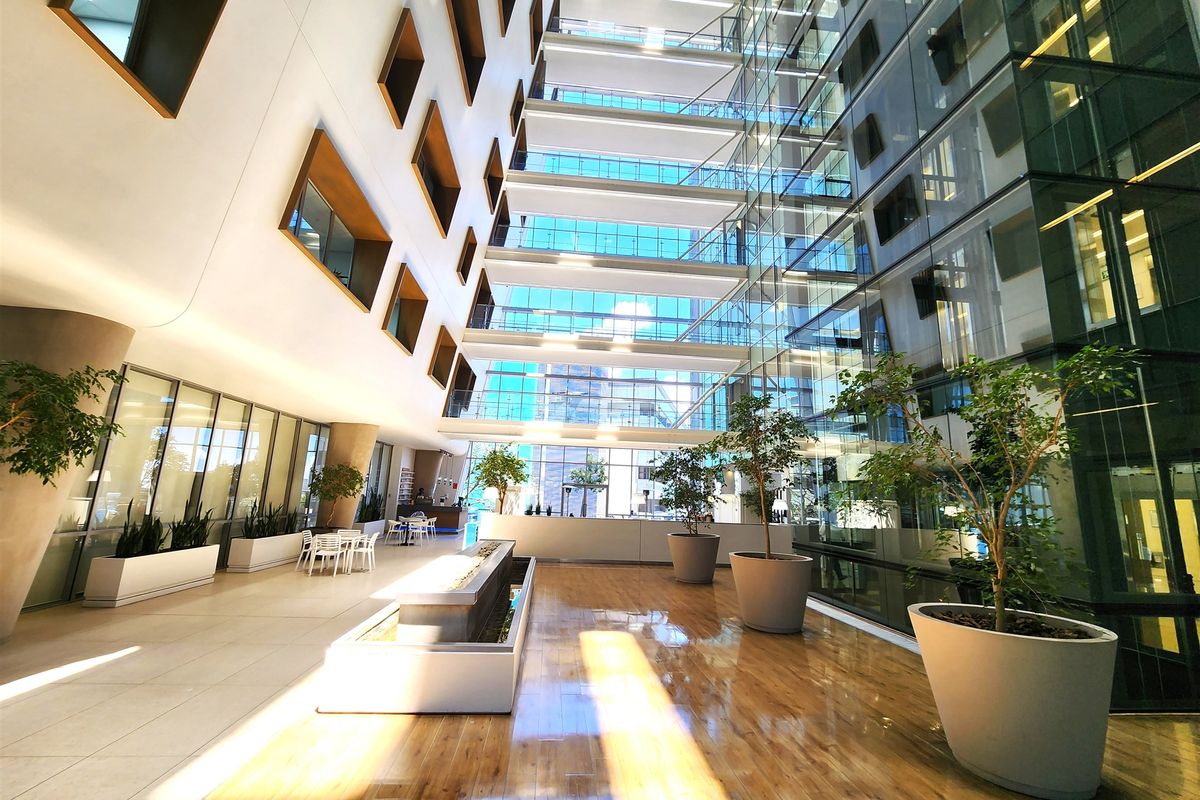 A modern, spacious office atrium featuring a multi-story glass facade, polished hardwood and tile flooring, and a central water feature.