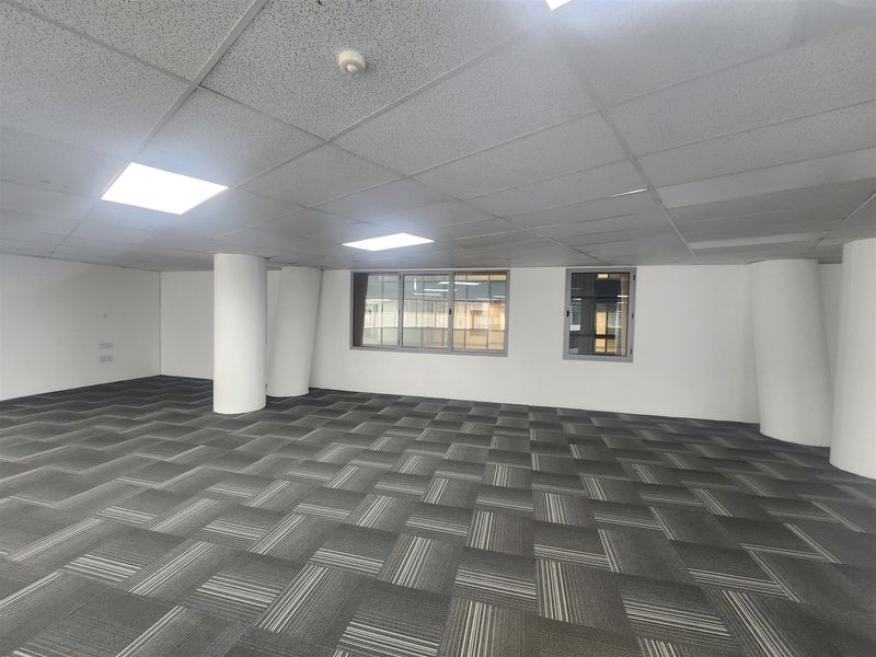 A spacious, empty open-plan office area featuring neutral carpet tiles, a suspended ceiling with recessed lighting, and large windows providing natural light.