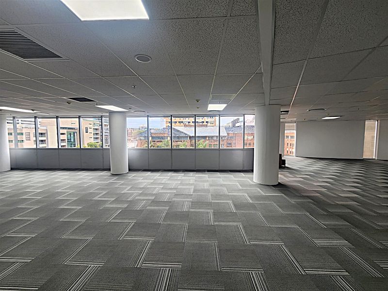 Spacious open-plan office area featuring large windows with city views, carpeted flooring, and a suspended ceiling with integrated lighting.