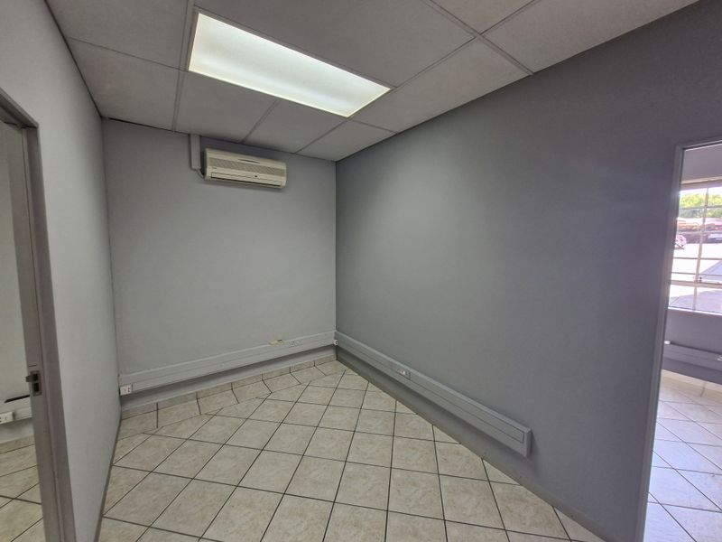 A clean, empty office space featuring tiled flooring, a drop ceiling with recessed lighting, and a wall-mounted air conditioning unit.