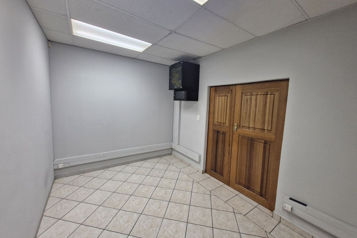 A clean, empty office space featuring tiled flooring, a suspended ceiling with recessed lighting, and a wall-mounted server rack.