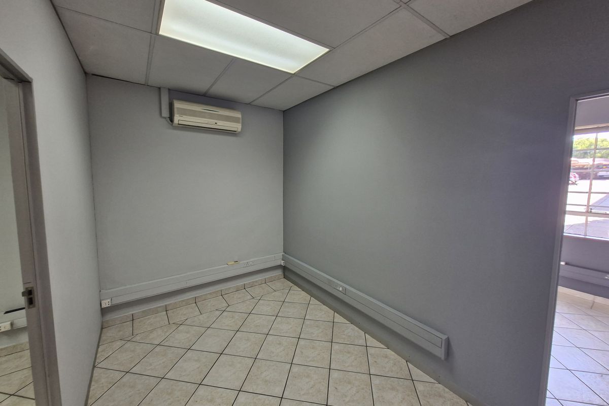 A clean, empty office space featuring tiled flooring, a drop ceiling with recessed lighting, and a wall-mounted air conditioning unit.
