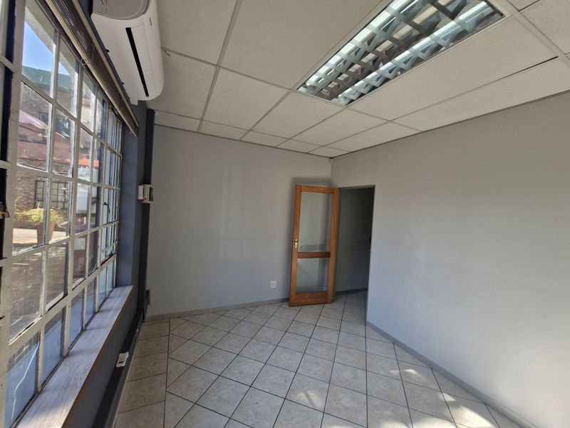 A vacant, well-lit office space featuring tiled flooring, a suspended ceiling with recessed lighting, and a split-system air conditioning unit.