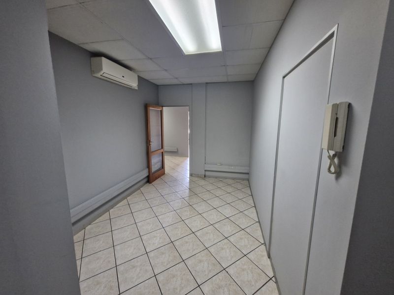 A vacant commercial office space featuring tiled flooring, a drop ceiling with integrated lighting, and a wall-mounted air conditioning unit.