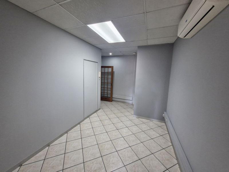 A clean, empty commercial hallway featuring tiled flooring, a suspended ceiling with recessed lighting, and a wall-mounted air conditioning unit.