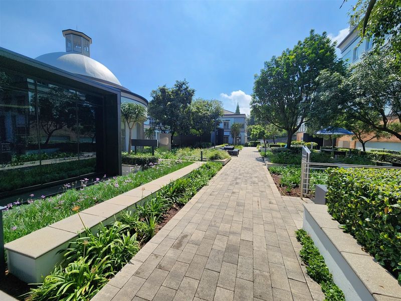A well-maintained outdoor courtyard area featuring a paved walkway, landscaped garden beds, and modern architectural elements including a glass facade and domed roof.
