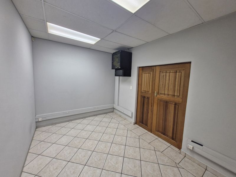 A clean, empty office space featuring tiled flooring, a suspended ceiling with recessed lighting, and a wall-mounted server rack.
