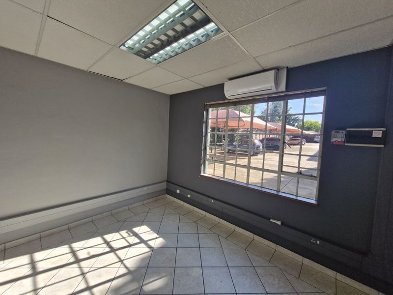 A clean, empty office space featuring tiled flooring, a suspended ceiling with recessed lighting, a split-system air conditioner, and security-barred windows.