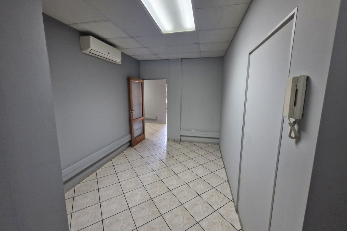 A vacant commercial office space featuring tiled flooring, a drop ceiling with integrated lighting, and a wall-mounted air conditioning unit.