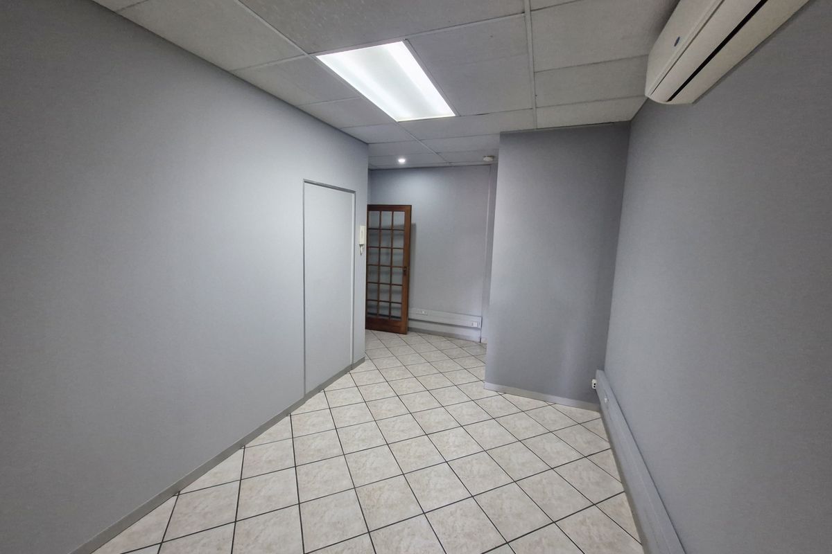 A clean, empty commercial hallway featuring tiled flooring, a suspended ceiling with recessed lighting, and a wall-mounted air conditioning unit.